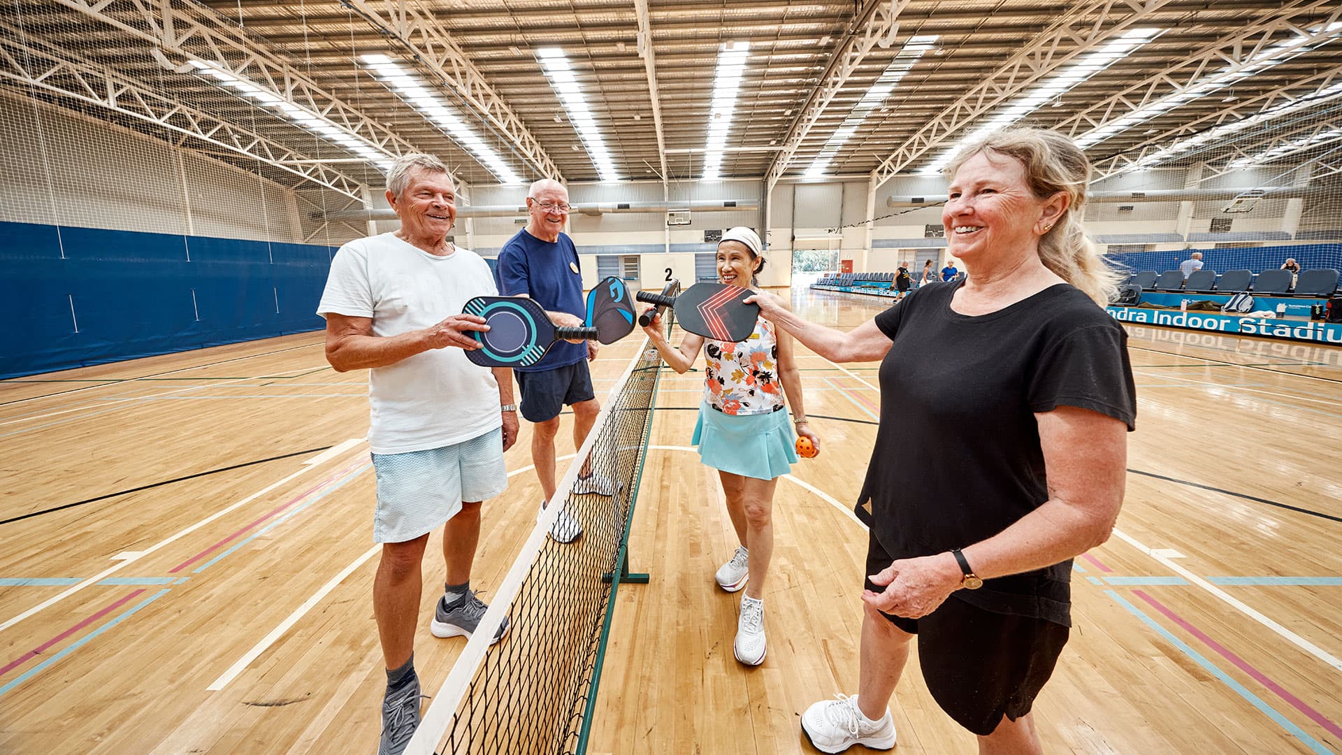 Four people stand on an indoor court holding pickleball paddles near a net