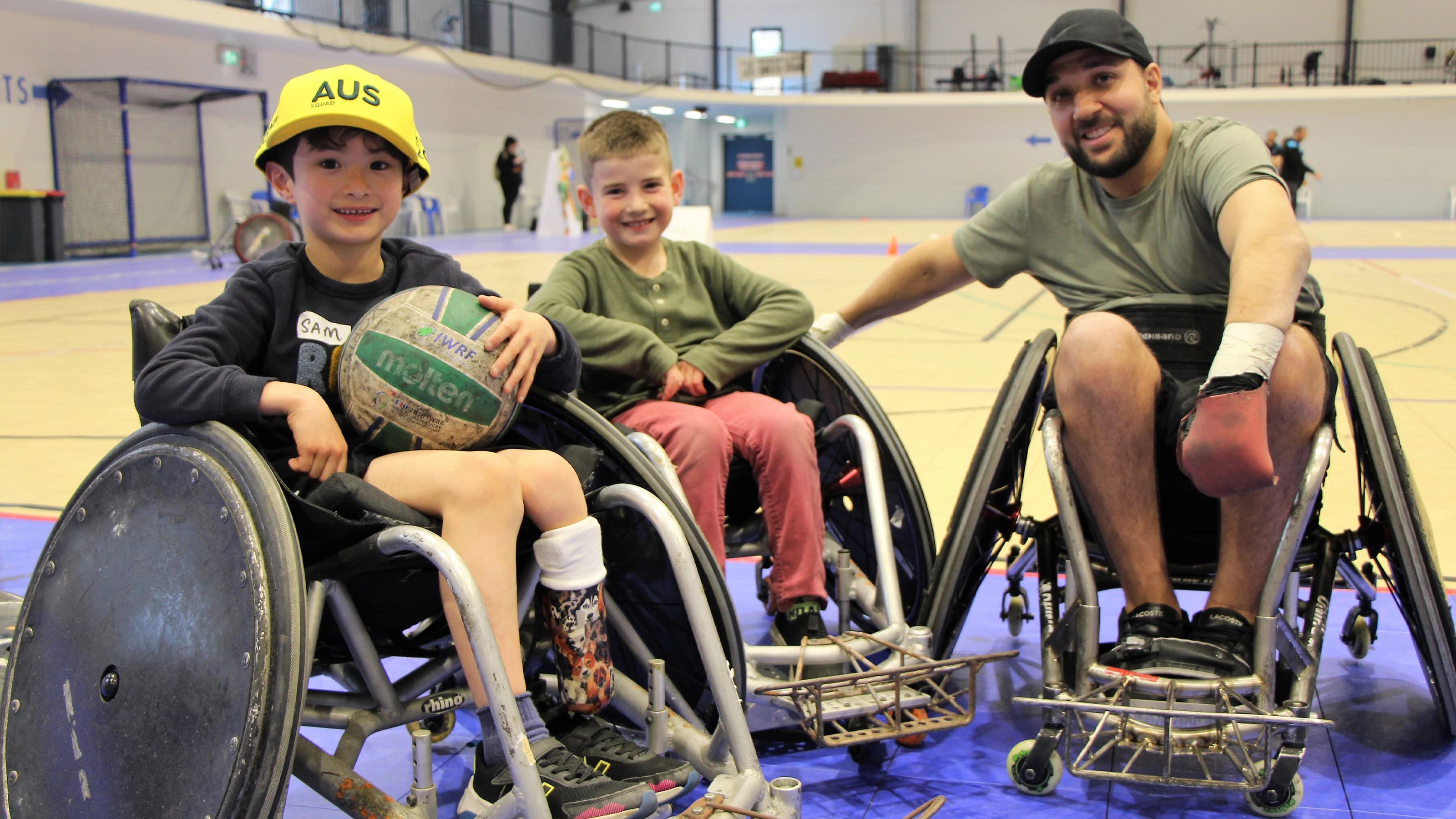 Two children and one adult in sport wheelchairs on an indoor sports court